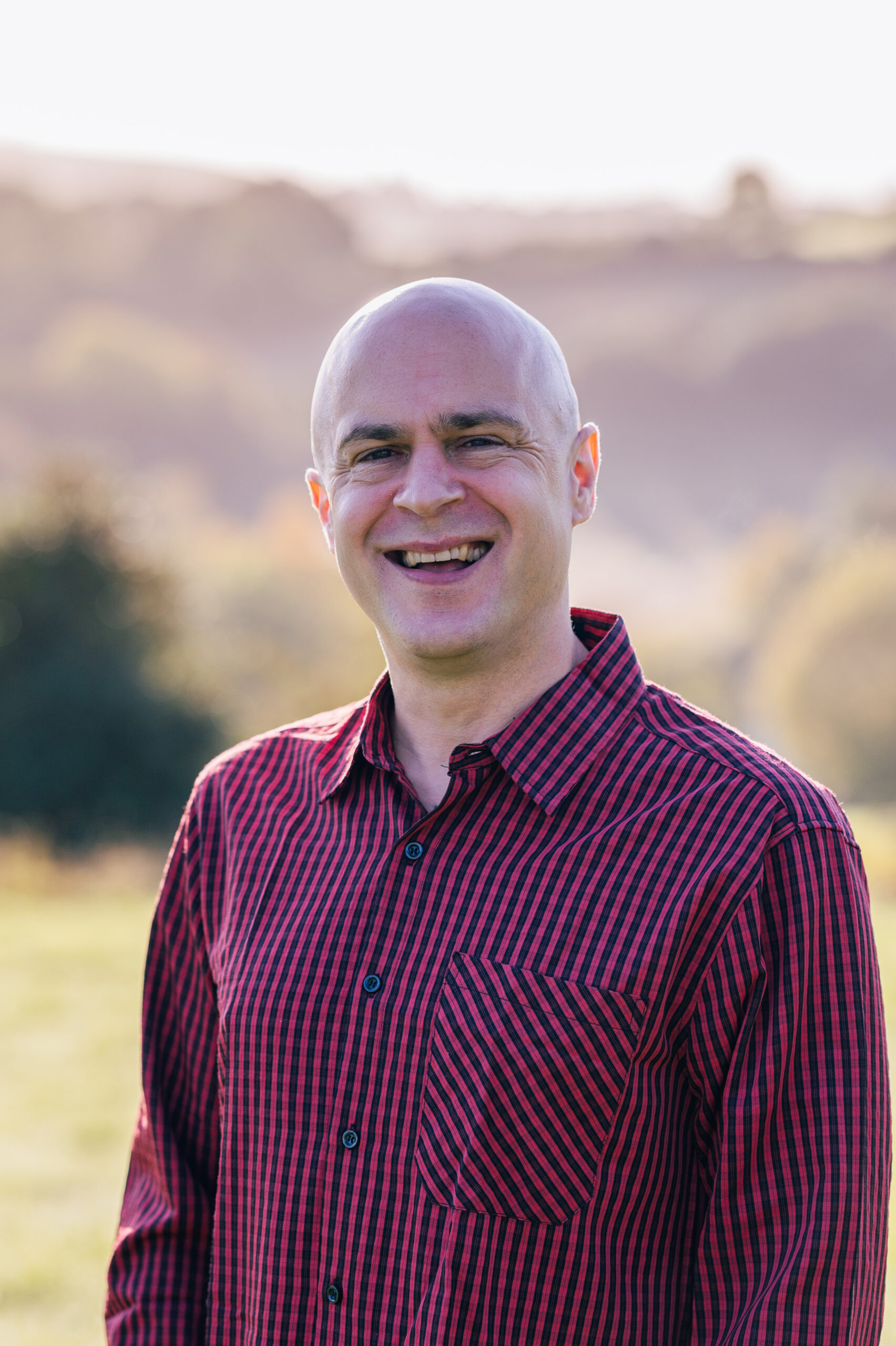 Andy Killworth in a field. Andy wears a red and black checked shirt and is smiling. In the background we can vaguely see hlls and field