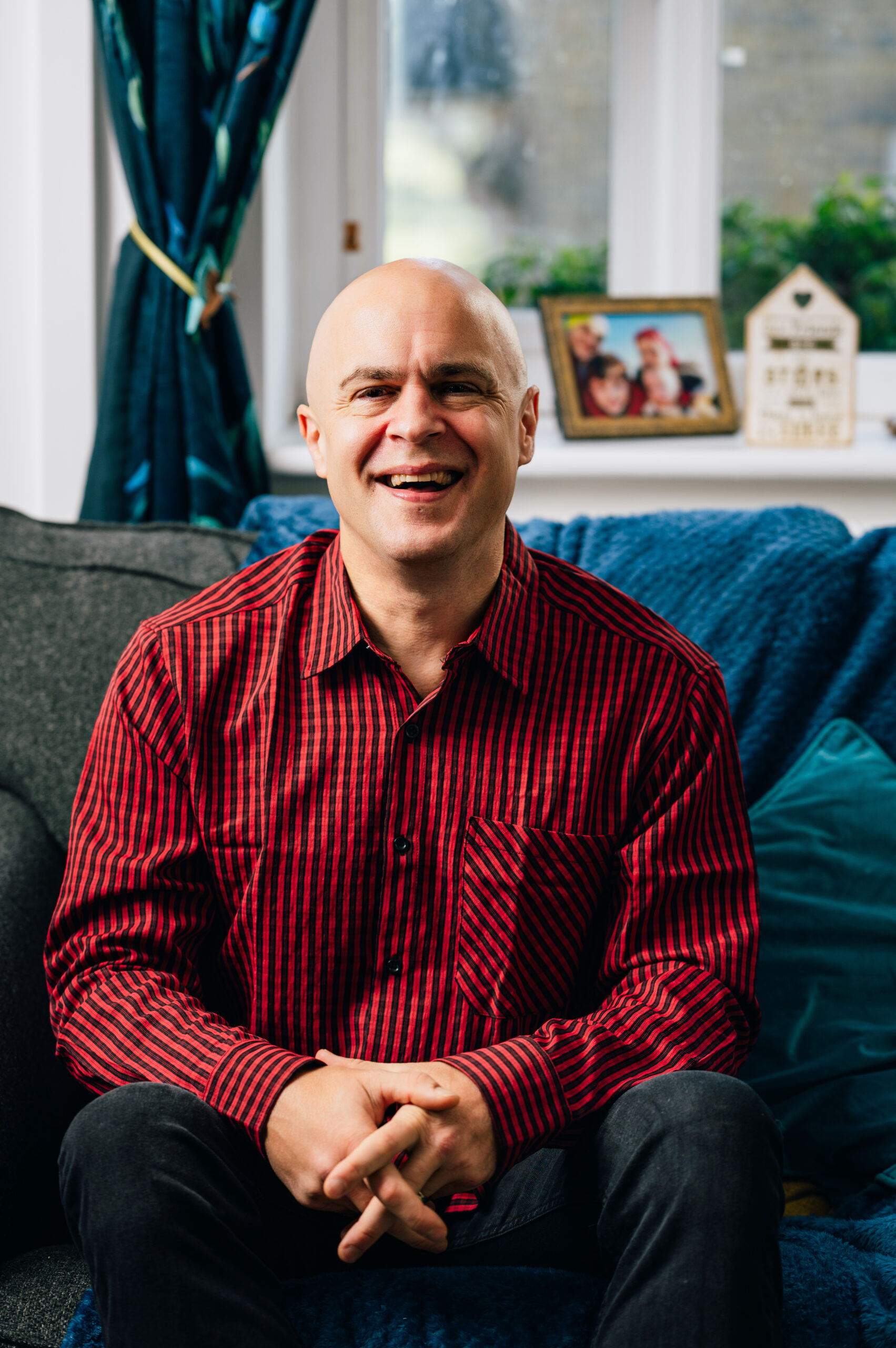 A photo of Andy Killworth. He's sat down on a sofa, wearing a red and black shite and black jeans. He's smiling. IN the background is a windowsill, window and green curtains.