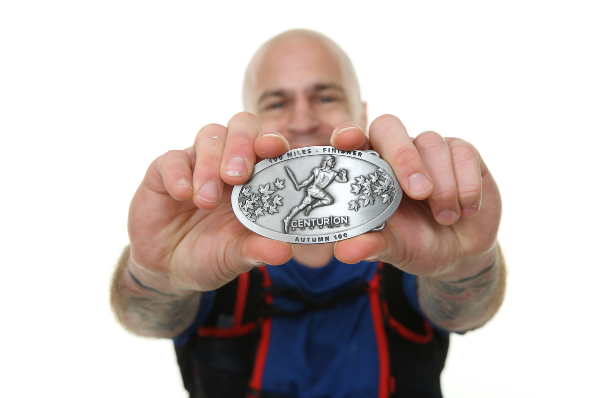 Andy Killworth holding a Centurion 100 Mile finisher buckle close to the camera. Andy is in the background with the camera's focus on the silver colored buckle from the 'Autumn 100' event. Andy is pictured on a white background, wearing a blue tshirt and black/red race best.