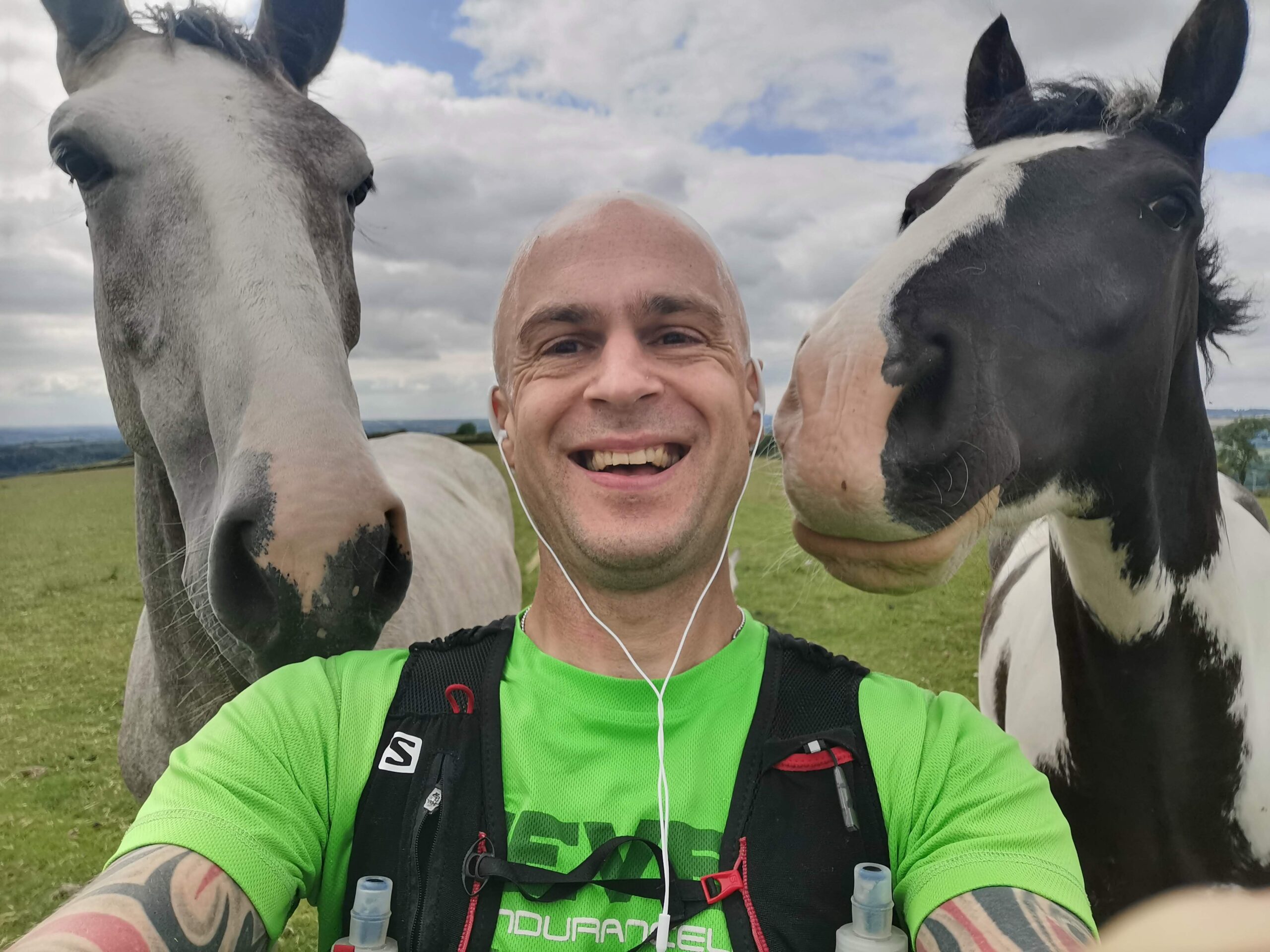 Andy Killworth in a selfie with two horses. Andy is in the centre, smiling broadly, wearing a green running shirt and black running vest. There is one horse each side of him (we see the horses from shoulder height upwards). The horse to the left is grey and white, the horse to the right is black and white.
