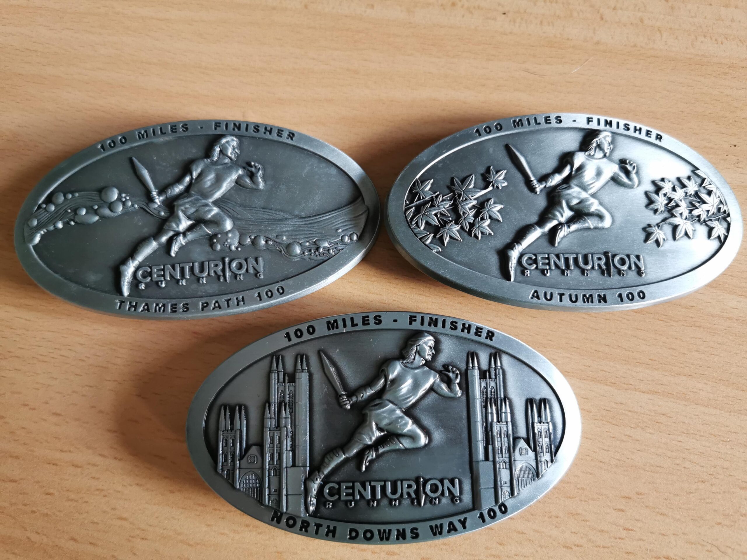 Three Centurion 100 mile ultra marathon buckles. They are pictured on a beige background. The buckles are silver coloured and all say '100 Miles - Finisher'. They each depict a centurion, with other images relevant to that race. The buckles are for Thames Path 100, Autumn 100 and North Downs 100.