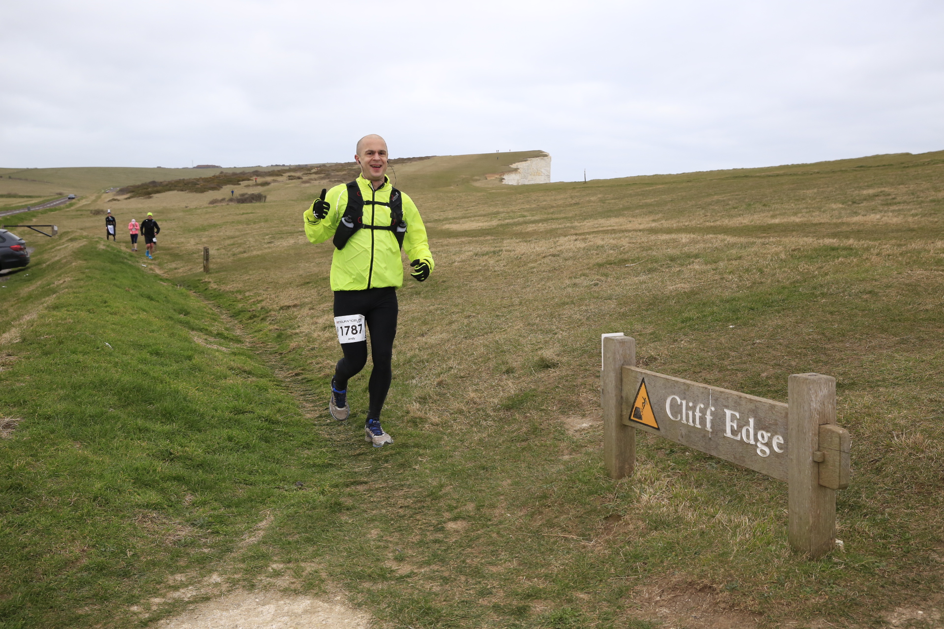 Andy Killworth running the CTS Endurance Life Dorset ultra marathon. Andy is pictured near the cliffs. There's a sign in the foreground that says 'Cliff Edge'. Andy is running along the path amongst short grass. He's wearing a bright yellow jacket, black gloves, black race  vest and black running tights. He's smiling and making a 'thumbs up' gesture with his right hand. Three other runners are in the background.