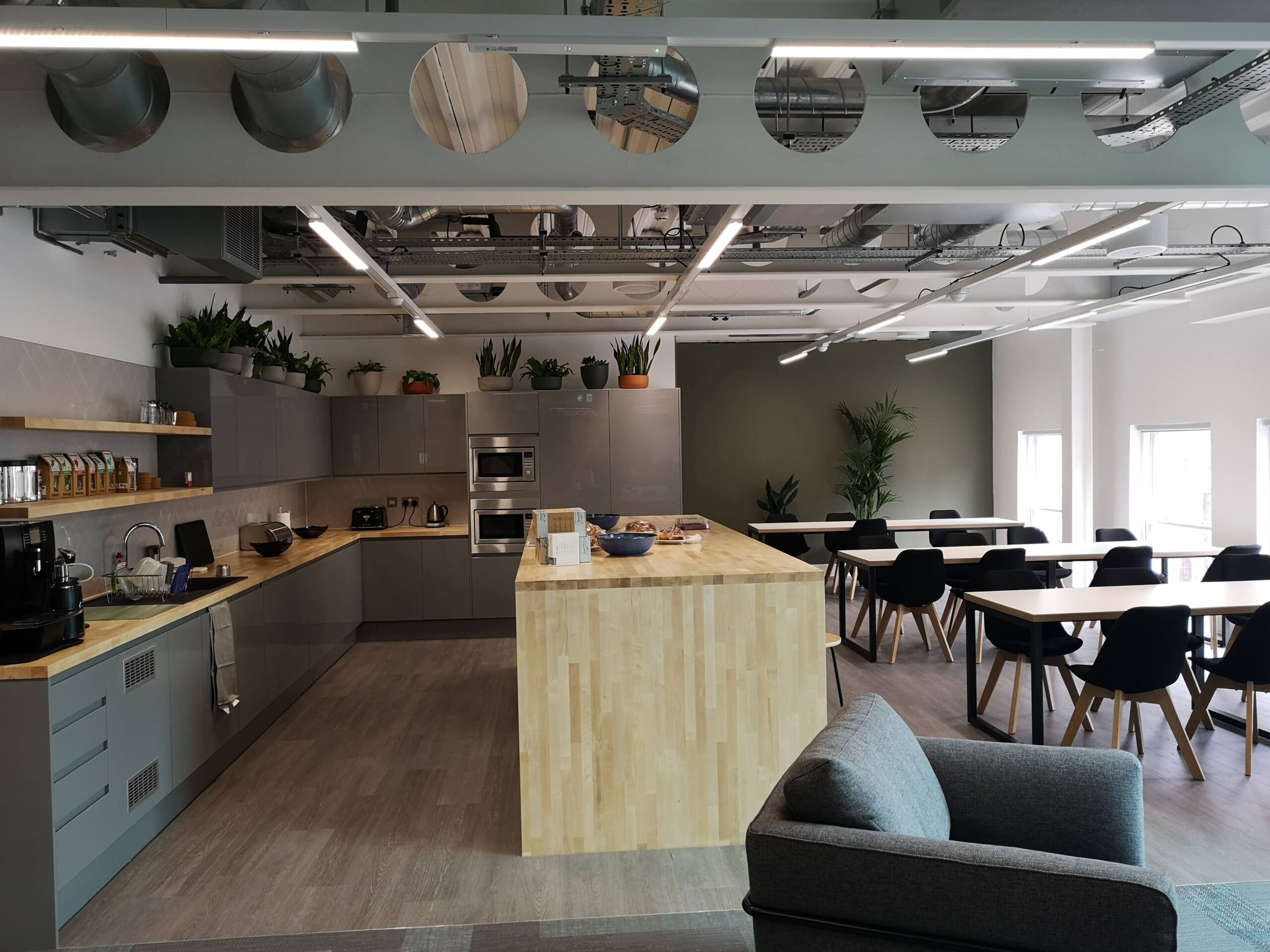 The kitchen and dining area at the old iTech Media office in Camden London. Photo shows a contemporary grey and light wood theme. We see kitchen units, a dining area with chairs, and a grey armchair in the foeground. Towards the rear and on top of the kitchen units are green plants