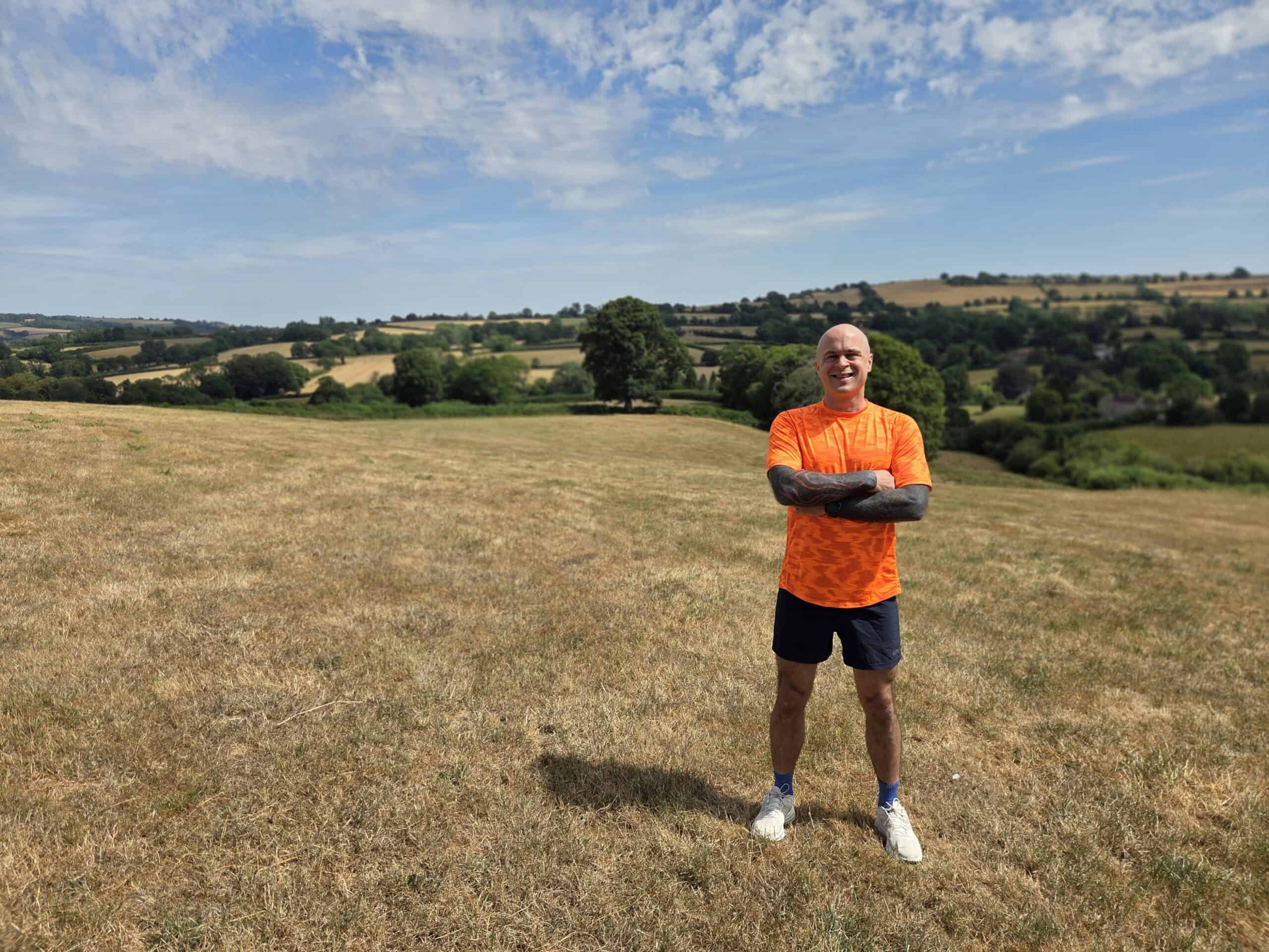 Andy Killworth Andy Killworth standing on a hill wearing a bright orange tshirt, blue running shorts and white running shoes. He has his arms crossed and we see both arms have 'black out' tattoos. He's stood on a grass hill with rolling hills and blue sky in the background.